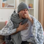 Zakażenia RSV: brak leczenia antywirusowego mężczyzna siedzi na kanapie, obwiązany szalikiem, w czapce i pod kocem. kaszle łapiąc się za pierś