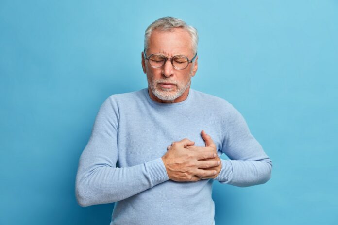 Senior man in spectacles presses hand to chest has heart attack suffers from unbearable pain closes eyes wears optical glasses poses against blue background. People age and problems with health starszy mężczyzna w niebieskim swetrze łapie się za serce