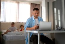 Rodzice coraz częściej sięgają po „dr Google”. Internet – wsparcie czy pułapka? na pierwszym planie mężczyzna siedzi przy stole na którym leży laptop i sprawdza coś w komórce. na drugim planie dziewczynka i chłopiec leżą na łóżku