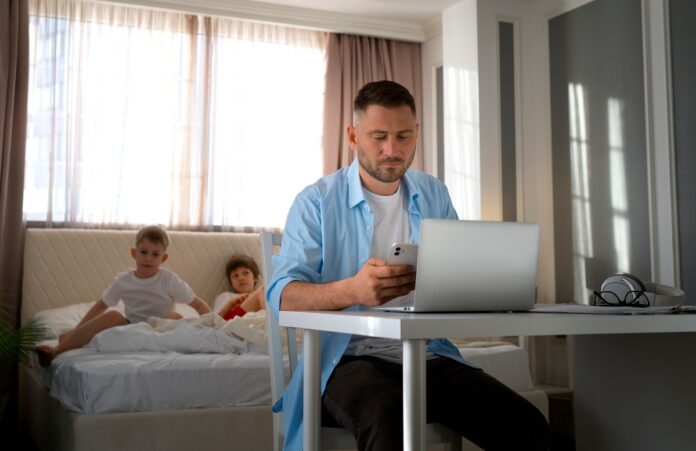 na pierwszym planie mężczyzna siedzi przy stole na którym leży laptop i sprawdza coś w komórce. na drugim planie dziewczynka i chłopiec leżą na łóżku