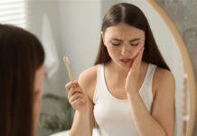 Tylko 1% Polaków nie ma problemów z dziąsłami młoda dziewczyna przed lustrem w łazience łapie się za policzek, w ręce ma szczoteczkę do zębów. zdjęcie ilustrujące ból zębów