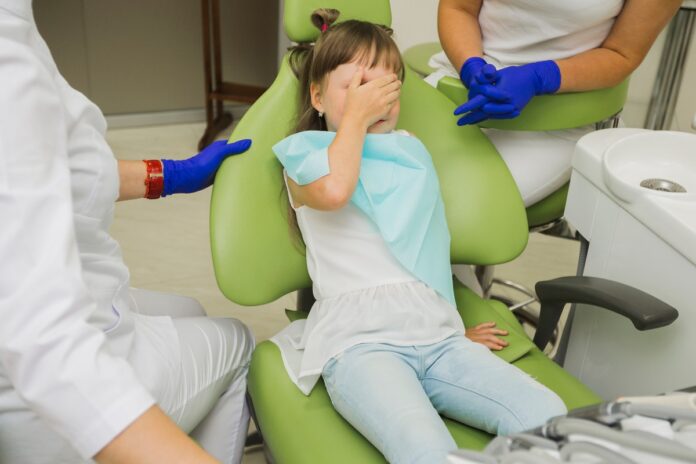 mała dziewczynka siedzi na fotelu dentystycznym i zakrywa oczy ze strachu