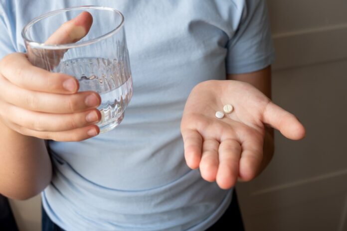 Pill and glass with water in the hand of child. Healthcare from pharmacy. Kid sit at home because of illness.